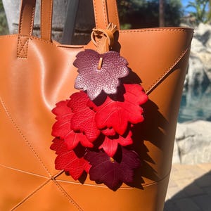 May include: A tan leather tote bag featuring a decorative hanging charm composed of multiple layered leaf-shaped cutouts in shades of deep burgundy, bright red, and dark crimson, all attached to the bag handle with a thin leather cord.