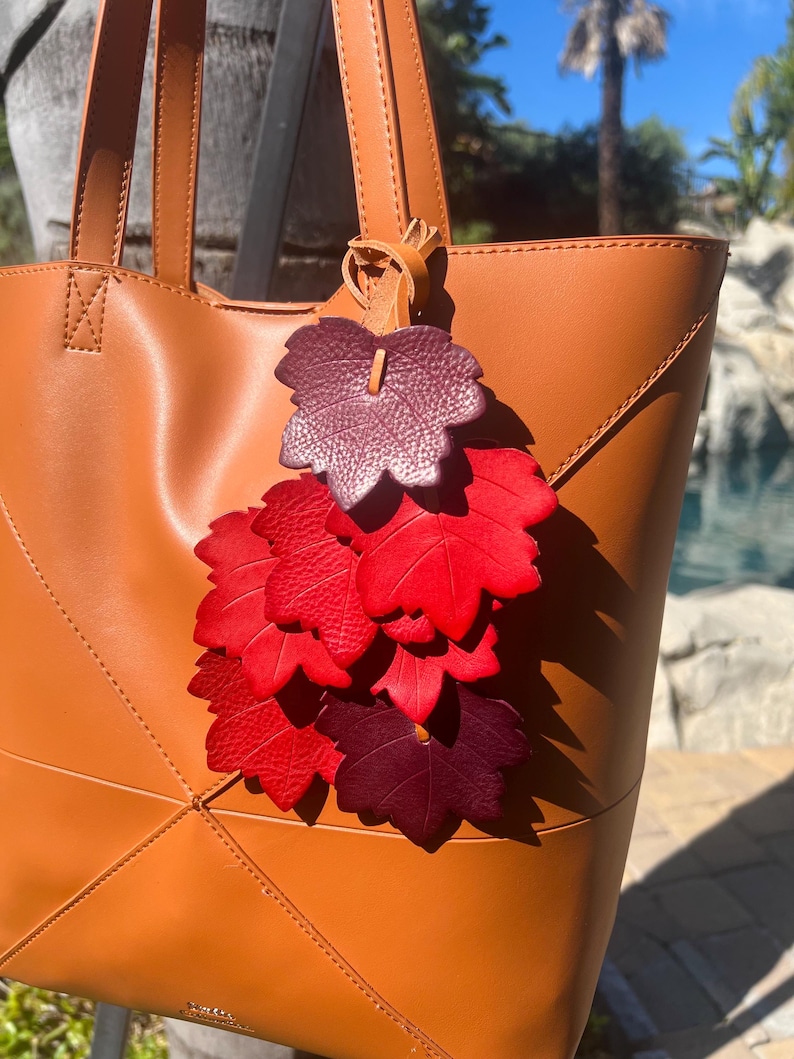 May include: A tan leather tote bag featuring a decorative hanging charm composed of multiple layered leaf-shaped cutouts in shades of deep burgundy, bright red, and dark crimson, all attached to the bag handle with a thin leather cord.