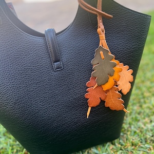 May include: A black textured leather handbag featuring a central vertical strap closure and a decorative leather charm attached to the handle. The charm consists of multiple oak leaf-shaped cutouts in shades of olive green, orange, and brown.