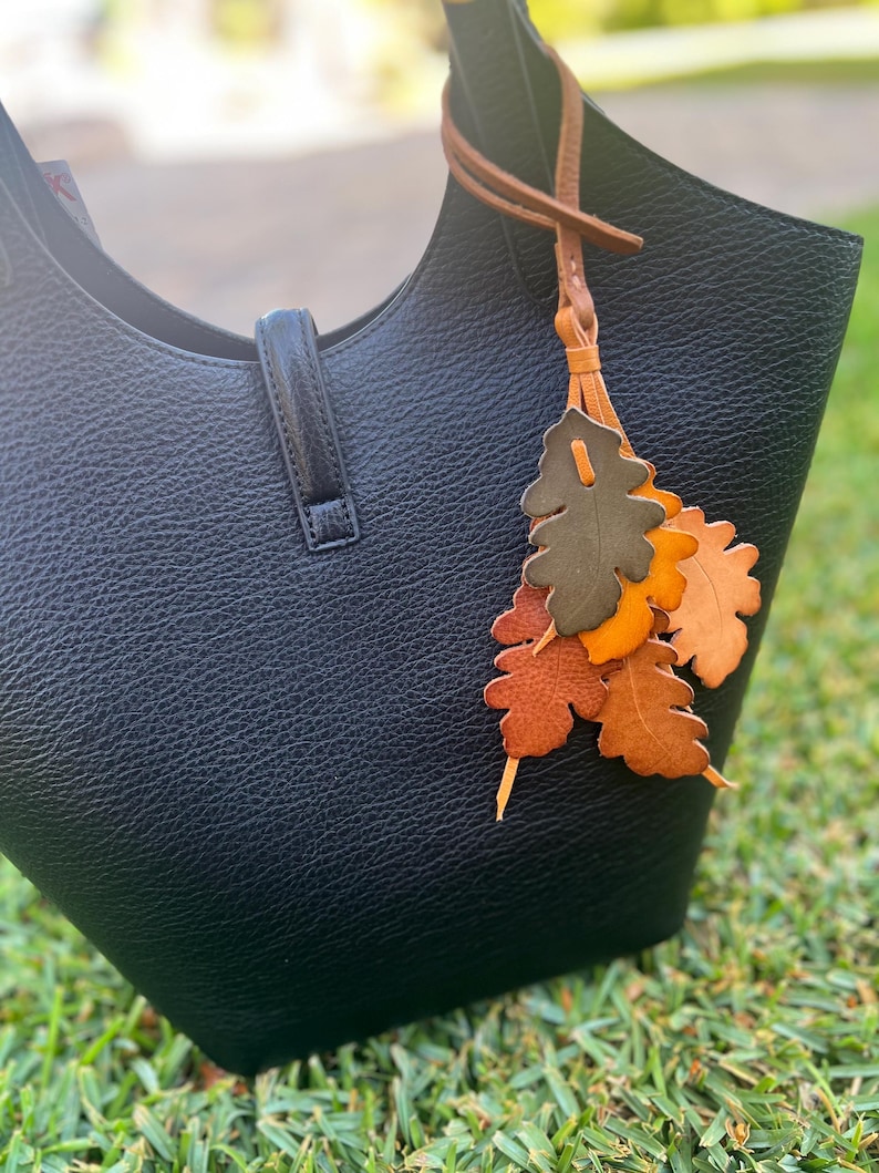 May include: A black textured leather handbag featuring a central vertical strap closure and a decorative leather charm attached to the handle. The charm consists of multiple oak leaf-shaped cutouts in shades of olive green, orange, and brown.