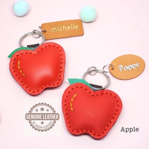 May include: Two red leather apple-shaped keychains with green leaves and yellow stitching. Each keychain has a silver ring and a small leather tag with a name. One tag says "Michelle" and the other says "Poppy". The word "Apple" is also visible.