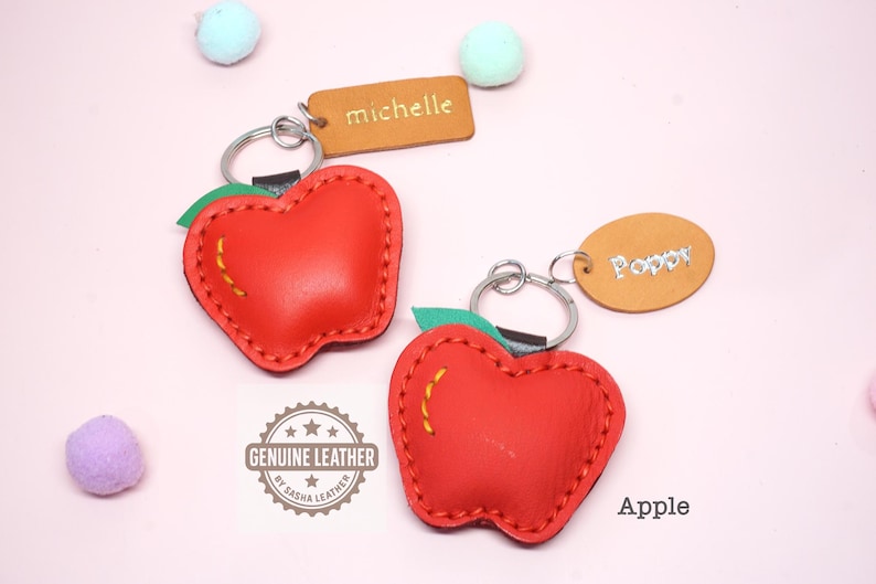 May include: Two red leather apple-shaped keychains with green leaves and yellow stitching. Each keychain has a silver ring and a small leather tag with a name. One tag says "Michelle" and the other says "Poppy". The word "Apple" is also visible.