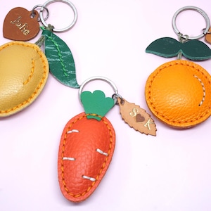May include: Leather keychains in the shape of a mango, orange, and carrot, each with a metal keyring. The mango is yellow, the orange is orange, and the carrot is red. Each keychain has a small leather tag with a name or initials.