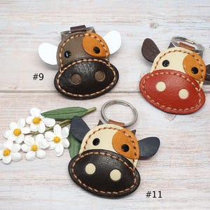 May include: Three leather cow-shaped keychains with metal rings. Keychain #9 is brown and tan with white ears. Keychain #10 is cream, orange, and dark brown. Keychain #11 is cream, orange, and dark brown with dark ears. White flowers are included.