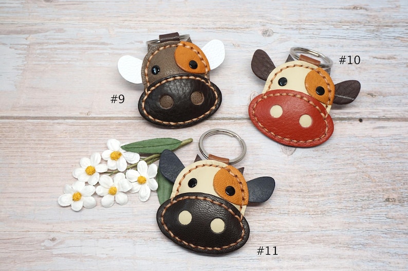May include: Three leather cow-shaped keychains with metal rings. Keychain #9 is brown and tan with white ears. Keychain #10 is cream, orange, and dark brown. Keychain #11 is cream, orange, and dark brown with dark ears. White flowers are included.