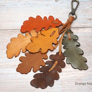 May include: A keychain featuring multiple oak leaf-shaped leather cutouts in shades of orange, yellow, brown, and olive green. The leaves are attached to a metal lobster clasp by thin leather strips. The text Orange keychain is visible at the bottom.