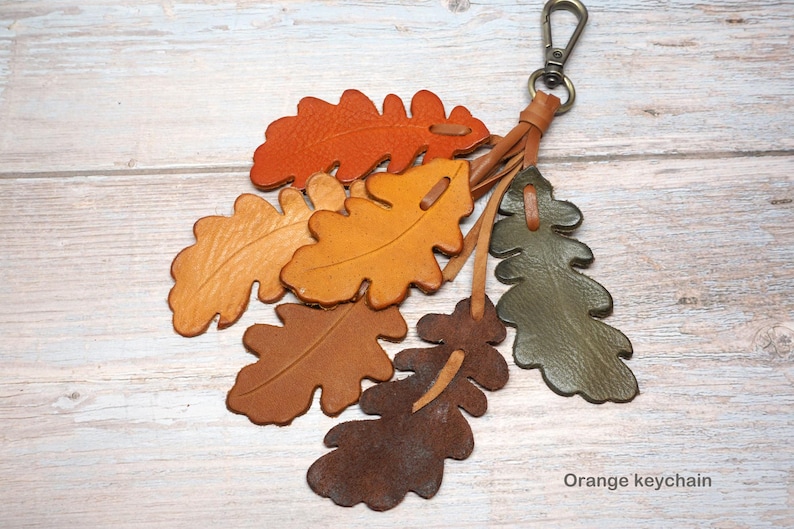 May include: A keychain featuring multiple oak leaf-shaped leather cutouts in shades of orange, yellow, brown, and olive green. The leaves are attached to a metal lobster clasp by thin leather strips. The text Orange keychain is visible at the bottom.