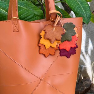 May include: A tan tote bag featuring two vertical handles and a decorative charm composed of multiple leather-like leaf cutouts in shades of brown, orange, yellow, green, red, and purple, attached to one of the bag's handle straps.