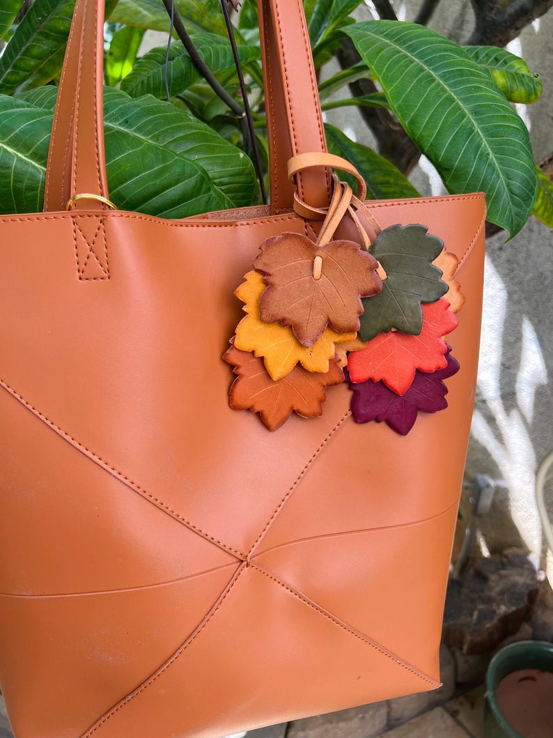 May include: A tan tote bag featuring two vertical handles and a decorative charm composed of multiple leather-like leaf cutouts in shades of brown, orange, yellow, green, red, and purple, attached to one of the bag's handle straps.