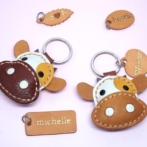 May include: Two cow-shaped leather keychains with white, tan, and dark brown patches, featuring metal rings. Additional leather tags include a leaf shape with S heart K, an oval with bestie, and a rectangular tag with the name michelle.