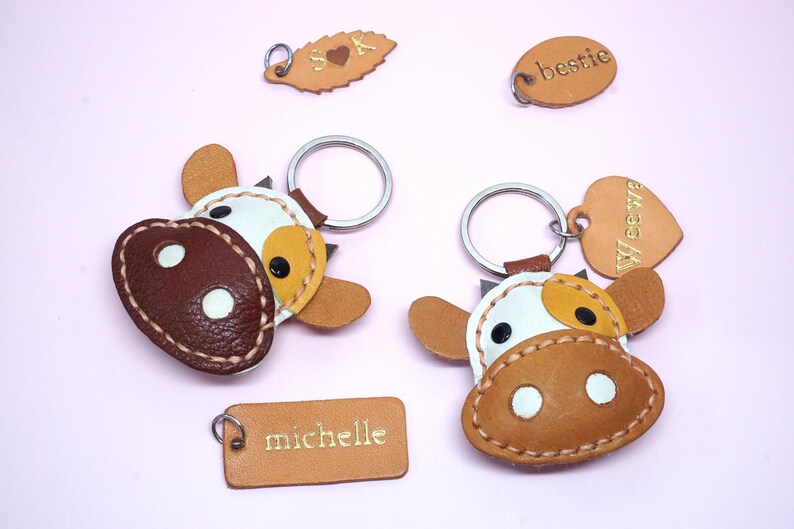 May include: Two cow-shaped leather keychains with white, tan, and dark brown patches, featuring metal rings. Additional leather tags include a leaf shape with S heart K, an oval with bestie, and a rectangular tag with the name michelle.