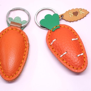 May include: Two orange leather carrot keychains with green tops and silver key rings. The carrots have yellow stitching around the edges and white stitching details. One keychain has a small leather tag with the initials "S" and "K" and a heart.