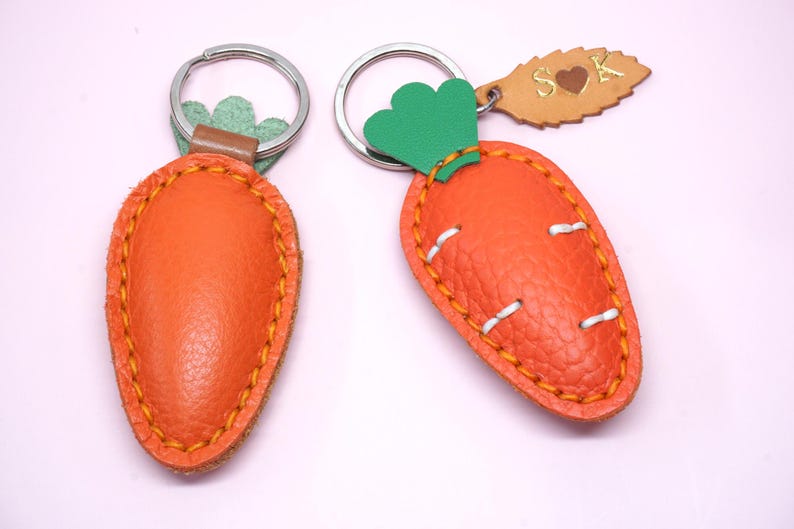 May include: Two orange leather carrot keychains with green tops and silver key rings. The carrots have yellow stitching around the edges and white stitching details. One keychain has a small leather tag with the initials "S" and "K" and a heart.