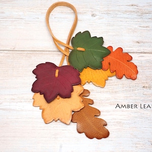May include: A decorative hanging ornament featuring a cluster of six leather leaves in shades of green, orange, yellow, burgundy, and tan, attached to a thin leather loop. The text AMBER LEAF is printed in black on the lower right side.