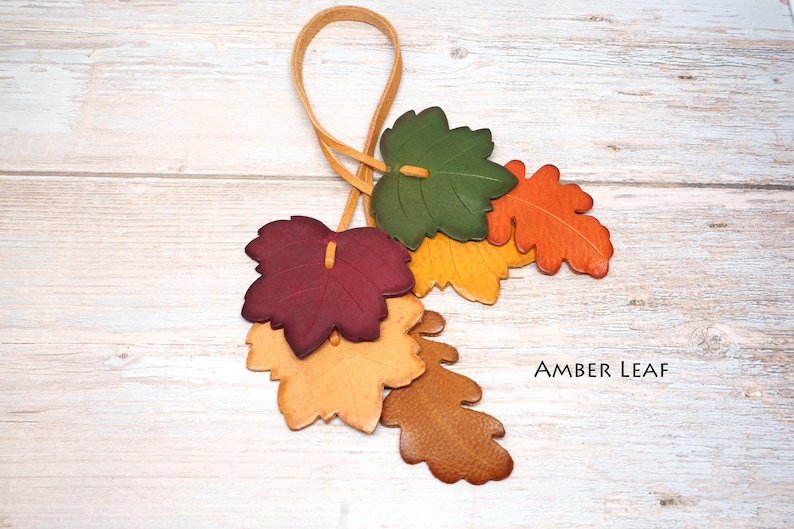 May include: A decorative hanging ornament featuring a cluster of six leather leaves in shades of green, orange, yellow, burgundy, and tan, attached to a thin leather loop. The text AMBER LEAF is printed in black on the lower right side.