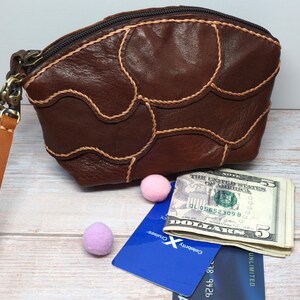 May include: A brown leather pouch with a zipper closure and a wrist strap. The pouch has a unique design with stitched accents. Beside it are a stack of US five-dollar bills, two credit cards, and two small felt balls.
