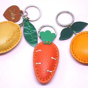 May include: A collection of leather keychains in the shapes of a mango, carrot, and orange. The mango is yellow, the carrot is orange, and the orange is a bright orange. Each keychain has a metal ring and is stitched with contrasting thread.
