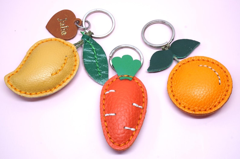 May include: A collection of leather keychains in the shapes of a mango, carrot, and orange. The mango is yellow, the carrot is orange, and the orange is a bright orange. Each keychain has a metal ring and is stitched with contrasting thread.