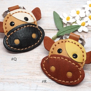 May include: Two cow-shaped leather keychains with metal rings. Keychain #Q features a tan and black face with black eyes and tan nostrils. Keychain #R features a tan and brown face with black eyes and tan nostrils. Both have visible stitching.