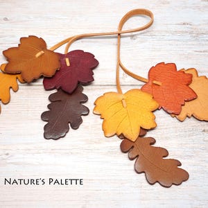 May include: A decorative hanging ornament featuring multiple leather-like leaves in shades of yellow, orange, brown, and deep red, suspended from thin tan cords. The text "NATURE'S PALETTE" is printed in black in the lower left corner of the image.