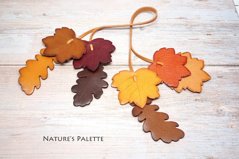 May include: A decorative hanging ornament featuring multiple leather-like leaves in shades of yellow, orange, brown, and deep red, suspended from thin tan cords. The text "NATURE'S PALETTE" is printed in black in the lower left corner of the image.