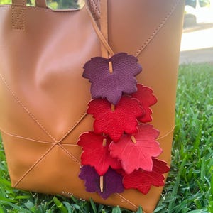 May include: A tan leather tote bag featuring a decorative hanging charm made of multiple textured leaf-shaped cutouts in shades of deep purple, bright red, and crimson, all attached to a thin leather cord tied to the bag handle.