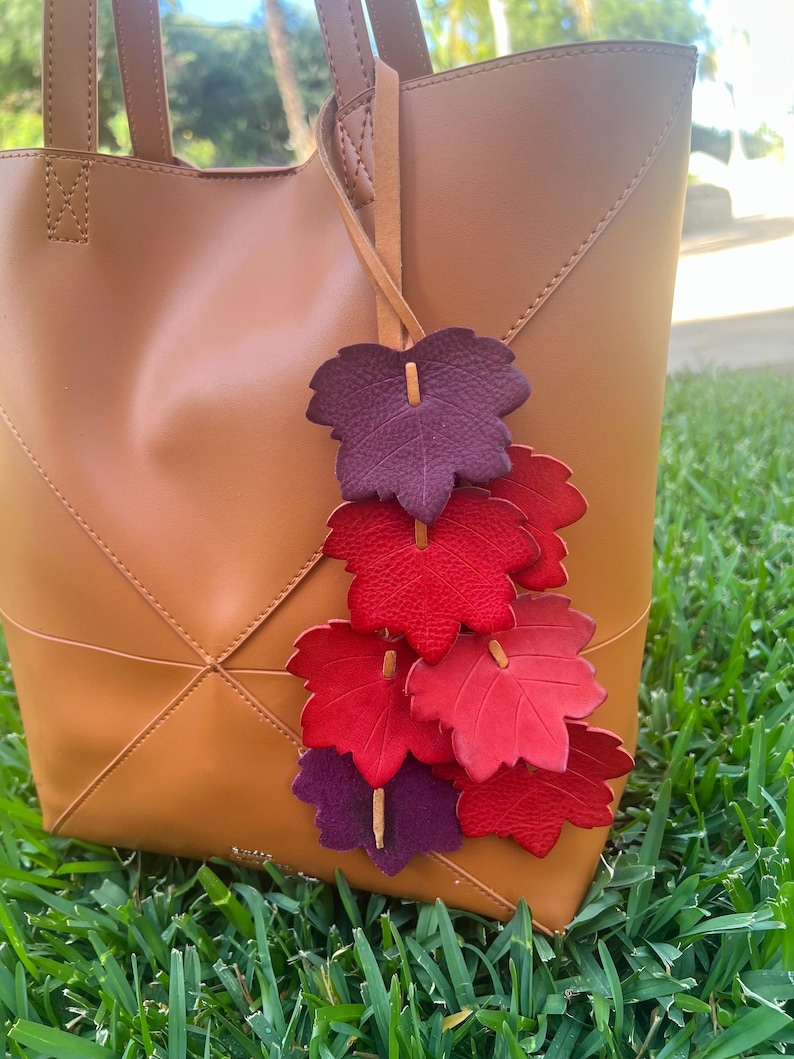 May include: A tan leather tote bag featuring a decorative hanging charm made of multiple textured leaf-shaped cutouts in shades of deep purple, bright red, and crimson, all attached to a thin leather cord tied to the bag handle.