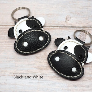 May include: Two cow-shaped keychains made of black and white textured material. Each keychain features a metal ring, stitched edges, and a stylized face with black and white patches. The text Black and White is printed below the keychains.