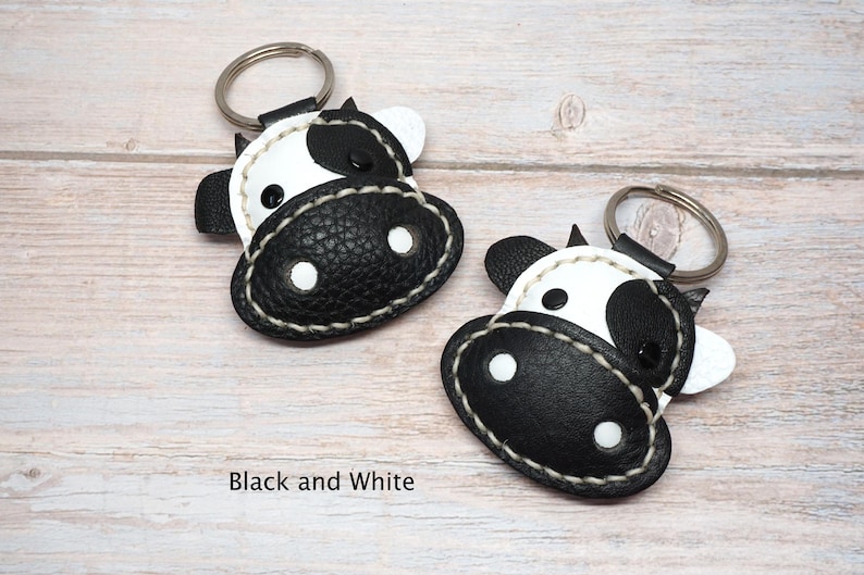 May include: Two cow-shaped keychains made of black and white textured material. Each keychain features a metal ring, stitched edges, and a stylized face with black and white patches. The text Black and White is printed below the keychains.