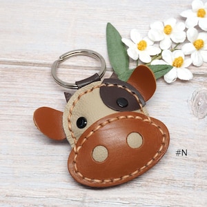 May include: A cow-shaped keychain crafted from beige and brown leather pieces stitched together with thick tan thread. The design features a dark brown patch over one eye, a large brown snout with two beige circles, and a silver metal ring.