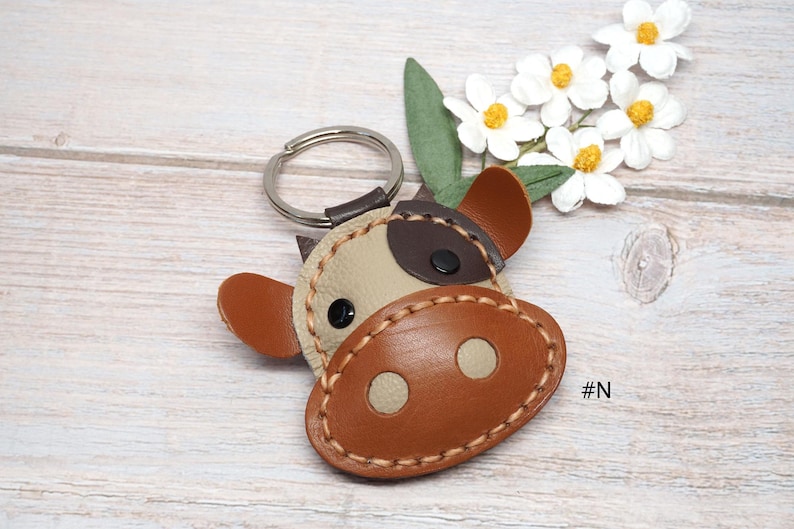 May include: A cow-shaped keychain crafted from beige and brown leather pieces stitched together with thick tan thread. The design features a dark brown patch over one eye, a large brown snout with two beige circles, and a silver metal ring.