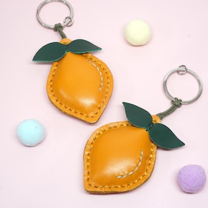 May include: Two lemon-shaped keychains with green leaves and silver key rings. The keychains are yellow with stitched details. Scattered around are three small, colorful, round objects on a pink background.