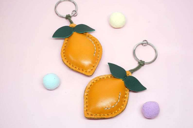 May include: Two lemon-shaped keychains with green leaves and silver key rings. The keychains are yellow with stitched details. Scattered around are three small, colorful, round objects on a pink background.