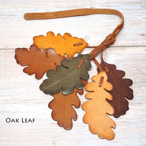 May include: A decorative hanging ornament featuring six individual oak leaf shapes crafted from leather in various shades of brown, tan, olive green, and yellow. The leaves are attached to a thin leather strap with a knotted loop at the top.