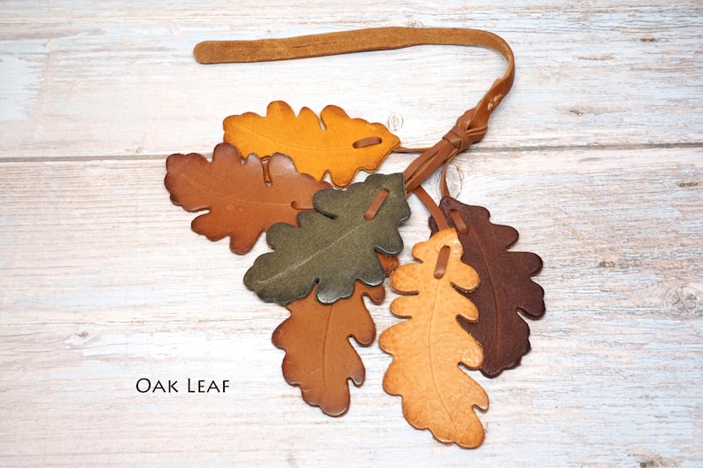 May include: A decorative hanging ornament featuring six individual oak leaf shapes crafted from leather in various shades of brown, tan, olive green, and yellow. The leaves are attached to a thin leather strap with a knotted loop at the top.