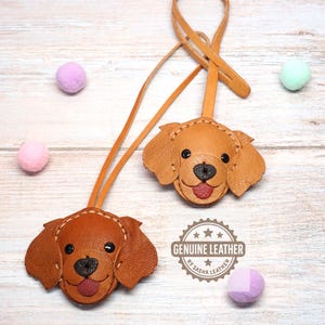 May include: Two tan leather dog-shaped keyrings with stitched details, black eyes, and red tongues. Each keyring has a leather strap. The image also includes a "Genuine Leather" stamp and several small, colourful pom-poms.