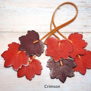 May include: A decorative hanging ornament featuring multiple leather leaves in shades of deep red, burgundy, and brown. The leaves are attached to a thin, light brown leather cord loop. The word Crimson is printed at the bottom of the image.