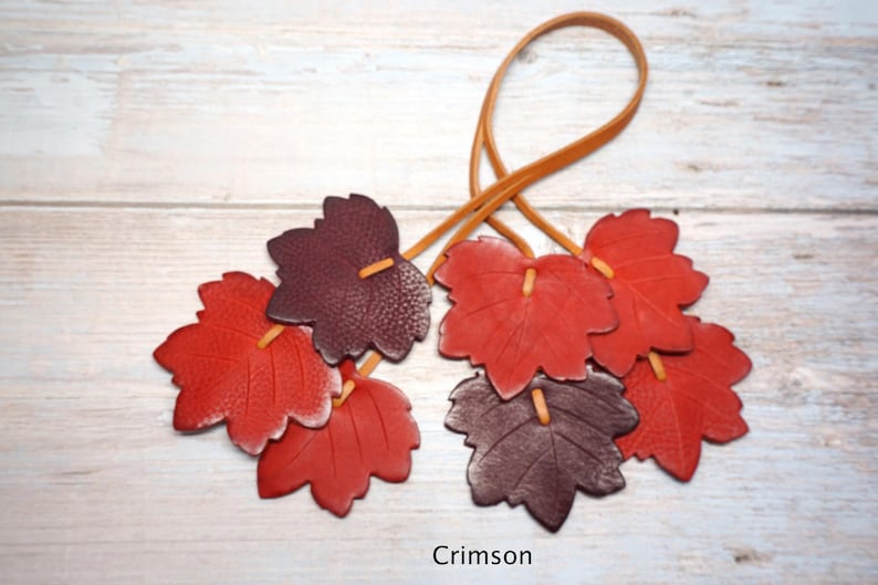 May include: A decorative hanging ornament featuring multiple leather leaves in shades of deep red, burgundy, and brown. The leaves are attached to a thin, light brown leather cord loop. The word Crimson is printed at the bottom of the image.
