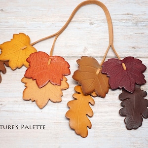 May include: A decorative hanging ornament featuring multiple leather leaves in shades of yellow, orange, brown, and deep red, suspended from a thin tan leather cord. The text NATURE'S PALETTE is printed in black in the lower left corner.