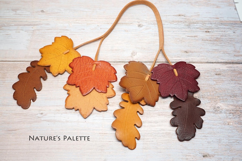 May include: A decorative hanging ornament featuring multiple leather leaves in shades of yellow, orange, brown, and deep red, suspended from a thin tan leather cord. The text NATURE'S PALETTE is printed in black in the lower left corner.