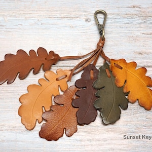 May include: A keychain featuring six leather oak leaf cutouts in shades of brown, tan, olive green, and yellow. The leaves are attached to a bronze-toned metal lobster clasp by thin leather cords. The text Sunset Keychain is printed in the corner.