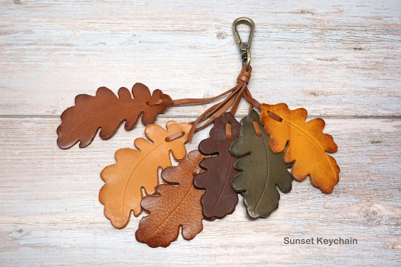 May include: A keychain featuring six leather oak leaf cutouts in shades of brown, tan, olive green, and yellow. The leaves are attached to a bronze-toned metal lobster clasp by thin leather cords. The text Sunset Keychain is printed in the corner.