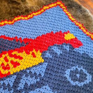 May include: A blue, red, and yellow crocheted baby blanket with a red and yellow truck design. The blanket has a scalloped edge.