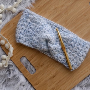 May include: A blue and white crocheted headband with a gold crochet hook on a wooden surface.