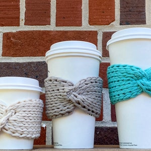 May include: Three white takeaway coffee cups with knitted cosy sleeves. The sleeves are in different colours: beige, grey, and teal.