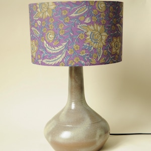 Könnte beinhalten: Eine Keramik-Tischlampe mit einem Lampenschirm mit Blumenmuster. Der Lampenschirm ist lila mit gelben und blauen Blumen. Der Fuß der Lampe ist hellbraun.