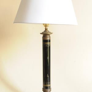Può includere: Una lampada da tavolo con paralume conico bianco su una base a colonna cilindrica nera con fasce decorative dorate. La lampada poggia su un piedistallo quadrato in marmo venato bianco e nero con piccoli piedini e un collo color ottone.