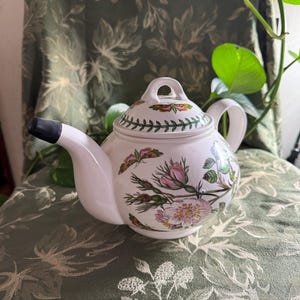 May include: White ceramic teapot with a black spout and handle. The teapot is decorated with pink roses, green leaves, and butterflies. The lid has a green and brown floral border. The teapot is sitting on a green floral patterned chair.