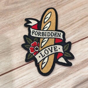 Op de afbeelding: Een geborduurde patch met een goudbruin stokbrood achter een crèmekleurig lint met de zwarte tekst FORBIDDEN LOVE. Het ontwerp bevat een rode bloem met groene bladeren en rode accenten, alles omlijnd met zwart garen voor een strakke afwerking.