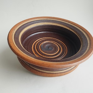 May include: A ceramic bowl with a spiral design in the center. The bowl has a wide rim with concentric bands of brown, tan, and beige. The interior is a dark brown with a spiral pattern.